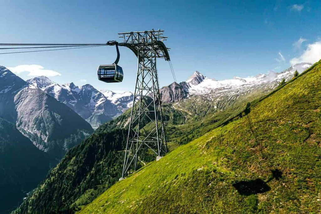 Seilbahnkabine über grünem Berghang mit schneebedeckten Gipfeln im Hintergrund