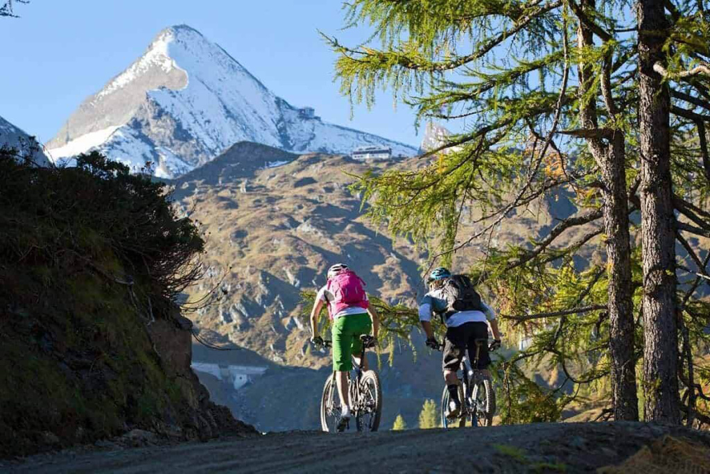 Zwei Radfahrer fahren auf einem Bergweg vor einem teils verschneiten Gipfel