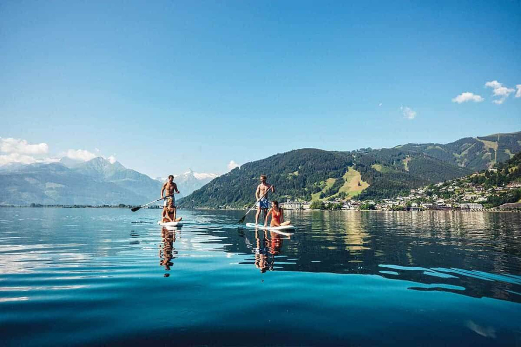 Vier Personen stehen auf SUP-Boards auf einem ruhigen See vor bewaldeten Bergen
