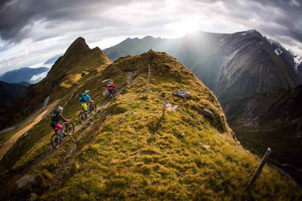 Drei Mountainbiker fahren auf einem schmalen Bergkamm bei dramatischer Bewölkung