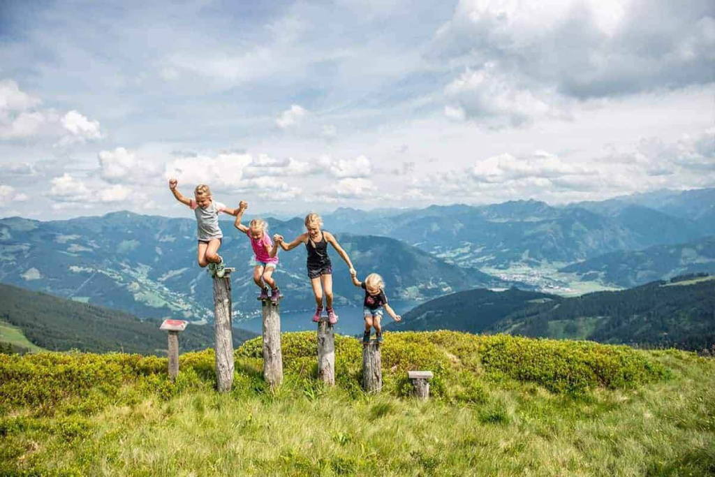 Vier Kinder springen auf Holzpfählen in einer alpinen Wiesenlandschaft mit weitem Talblick