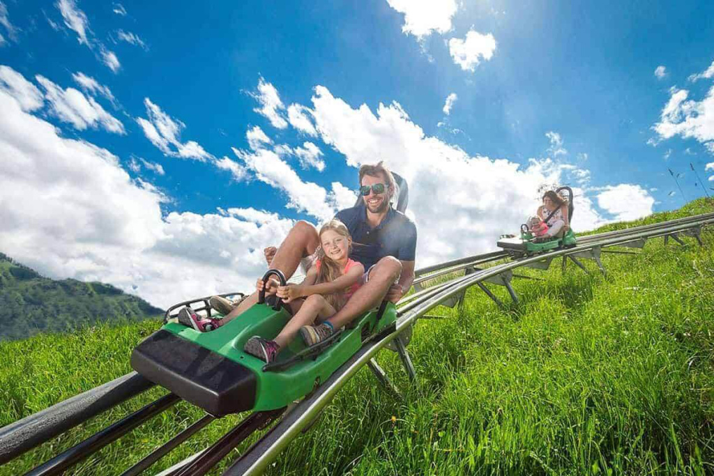 Zwei Personen fahren in grünen Sommerrodelbahnen über eine Wiese bergab