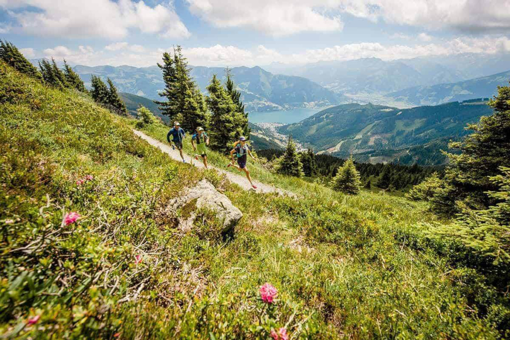 Drei Personen laufen auf einem Bergpfad über einer grünen Alm mit Blick auf einen See